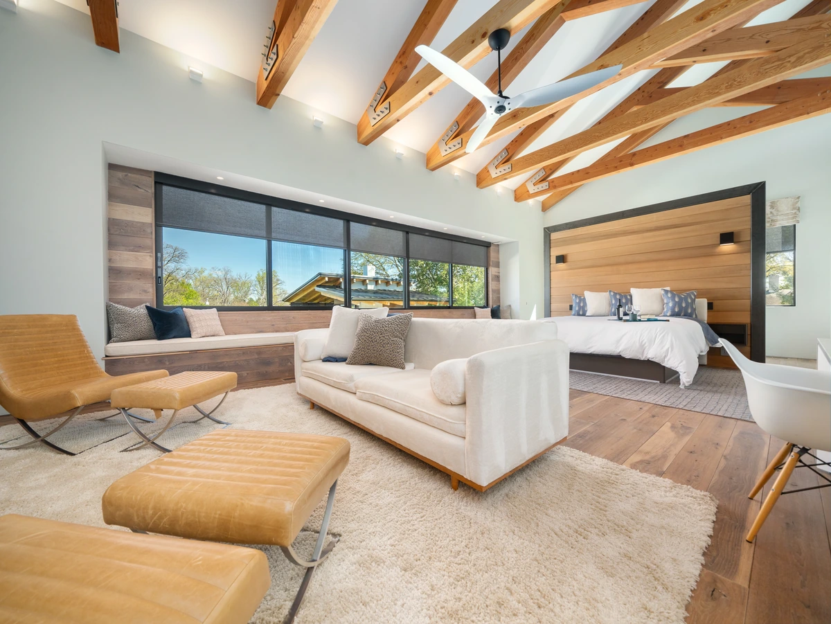 Bright luxury bedroom and sitting area with exposed wood beams, large windows, and a wood-paneled bed wall
